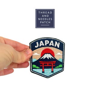 Könnte beinhalten: Gestickter Aufnäher mit einer japanischen Landschaft mit dem Fuji, einem roten Torii-Tor und dem Wort "JAPAN" in Weiß. Der Aufnäher hat einen marineblauen Rand und den Text "THREAD AND NEEDLES PATCH" in einem marineblauen Quadrat.