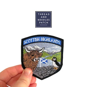 May include: Embroidered patch depicting a Highland cow, puffin, and Scottish flag, with the text "Scottish Highlands." The patch has a black border and a blue and white sky background. The patch is held by a hand.