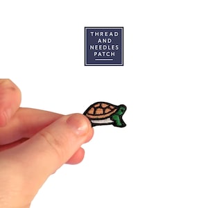 Könnte beinhalten: Ein kleiner, gestickter Schildkrötenaufnäher mit braunem Panzer, grünem Kopf und weißem Bauch. Der Aufnäher wird in einer Hand vor weißem Hintergrund gehalten. Der Text "THREAD AND NEEDLES PATCH" befindet sich in einem marineblauen Quadrat.