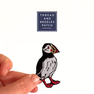 Op de afbeelding: Een geborduurde strijk-patch met een cartoon papegaaiduiker met een zwart-wit lichaam, rode snavel en poten en een gele oogring. De patch is gelabeld "THREAD AND NEEDLES PATCH".