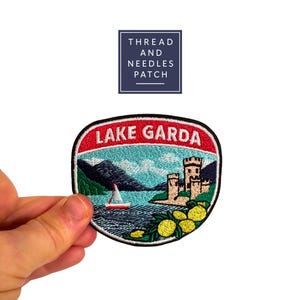 Könnte beinhalten: Gestickter Aufnäher mit einer Ansicht des Gardasees. Der Aufnäher hat einen roten Rand mit dem weißen Schriftzug "LAKE GARDA". Das Design umfasst ein Segelboot, Berge, eine Burg und Zitronen. Der Aufnäher wird von einer Hand gehalten.