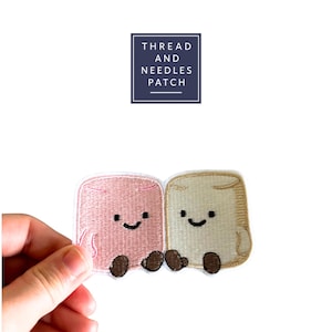 May include: Embroidered patch featuring two smiling marshmallow characters, one pink and one beige, with brown feet. The patch has the text "THREAD AND NEEDLES PATCH" in a navy blue square.