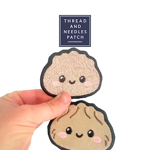May include: Two embroidered patches in the shape of dumplings with smiling faces and pink cheeks. The patches are tan with black outlines. The text "THREAD AND NEEDLES PATCH" is in a navy blue box above the patches.