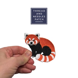 Pode incluir: Patch bordado com um panda-vermelho com pelo laranja, detalhes pretos e uma cauda listrada. O patch tem o texto "THREAD AND NEEDLES PATCH" em um retângulo azul marinho.