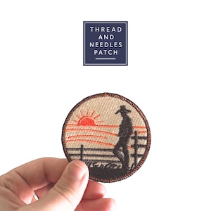 May include: Embroidered circular patch depicting a cowboy at sunset. The design features a red sun, orange horizon, and a fence. The patch has a beige background and a brown border. The text "THREAD AND NEEDLES PATCH" is in a navy blue square.