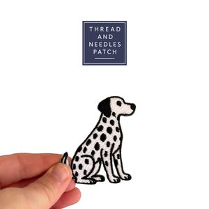 May include: An embroidered patch of a sitting Dalmatian dog with black spots on a white background. The patch has a black outline. The text "THREAD AND NEEDLES PATCH" is in a navy blue rectangle. The patch is held by a hand.