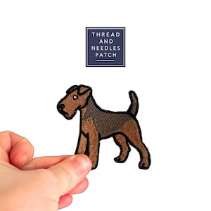 Könnte beinhalten: Gestickter Aufnäher mit einem braunen und schwarzen Airedale Terrier Hund. Der Aufnäher hat einen schwarzen Rand und befindet sich vor einem weißen Hintergrund. Der Text "THREAD AND NEEDLES PATCH" steht in einem marineblauen Quadrat.