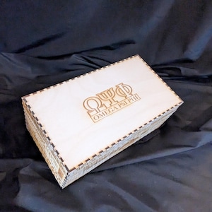 May include: A wooden box with a black and white checkered border. The top of the box is light brown and has the Greek letters "ΩΨΦ" and the text "OMEGA PSI PHI" engraved on it.