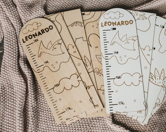 May include: A set of six wooden growth charts featuring dinosaur illustrations and the name "Leonardo". The charts are laid out in a fan shape on a pink knitted blanket with pom-poms.