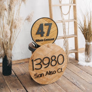 Może przedstawiać: Dwa okrągłe drewniane znaki z czarnymi numerami i tekstem. Górny znak zawiera napis "47 Alora Crescent", a dolny znak zawiera napis "13980 San Aliso Ct."