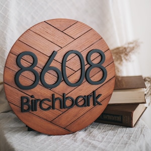 May include: A round wooden sign with a herringbone pattern. The sign has black numbers "8608" and the text "Birchbark" in black lettering.