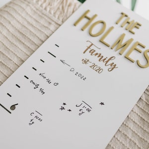 Puede incluir: Un gr&aacute;fico de crecimiento de madera blanco con letras doradas que dicen "THE HOLMES Family est 2020". El gr&aacute;fico tiene una l&iacute;nea de marcador negro con medidas de altura y fechas escritas en marcador negro.