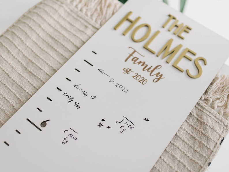 Puede incluir: Un gr&aacute;fico de crecimiento de madera blanco con letras doradas que dicen "THE HOLMES Family est 2020". El gr&aacute;fico tiene una l&iacute;nea de marcador negro con medidas de altura y fechas escritas en marcador negro.