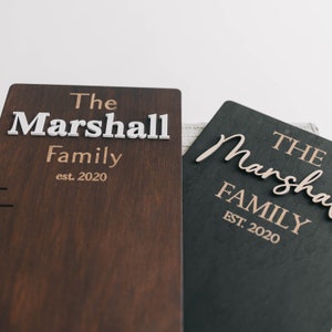 Puede incluir: Dos letreros de madera con el texto "The Marshall Family est. 2020" en diferentes fuentes y colores. El letrero de la izquierda es marr&oacute;n con letras blancas y el letrero de la derecha es negro con letras doradas.