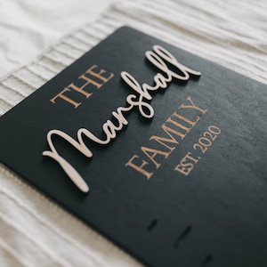 Puede incluir: Un letrero de madera negro con las palabras "The Marshall Family Est. 2020" en letras doradas. La palabra "Marshall" est&aacute; hecha de madera y est&aacute; unida al letrero.