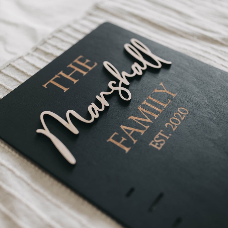 Puede incluir: Un letrero de madera negro con las palabras "The Marshall Family Est. 2020" en letras doradas. La palabra "Marshall" est&aacute; hecha de madera y est&aacute; unida al letrero.