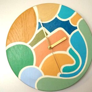 May include: A round wooden wall clock with a colorful abstract design. The clock face features a mix of colors including green, blue, orange, and beige. The clock hands are gold-colored.