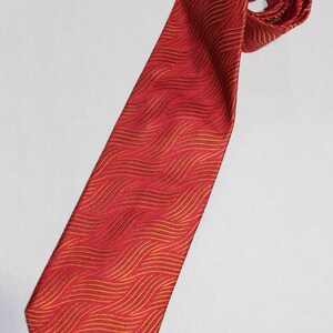 May include: A red tie with a repeating wavy pattern in shades of gold. The tie is displayed on a white background, with the end rolled up. The tie is a classic accessory for formal or business attire.