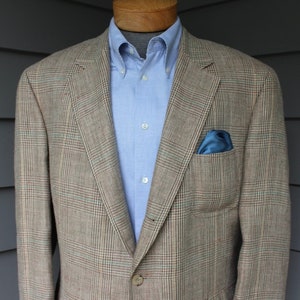 May include: A tan plaid blazer with a blue pocket square and a light blue button-down shirt. The blazer has a classic fit and is made of a textured fabric. The outfit is displayed on a wooden mannequin.