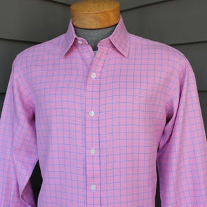 May include: Pink button-down shirt with a blue and red plaid pattern. The long-sleeved shirt has a classic collar and is buttoned up the front. The shirt is displayed on a wooden mannequin head.