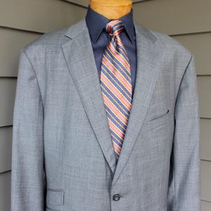 May include: A light gray suit jacket with a navy blue shirt and a striped tie. The tie features diagonal stripes in shades of pink, blue, and yellow. The jacket has a two-button closure and a chest pocket.