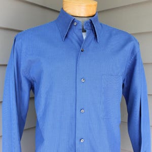 May include: A blue long-sleeve button-down shirt with a textured pattern. The shirt has a classic collar, a chest pocket, and is buttoned up. The shirt is displayed on a wooden mannequin head and shoulders.
