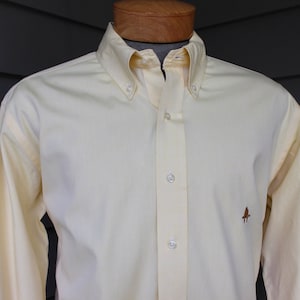 May include: A pale yellow long-sleeved button-down shirt with a small brown logo on the right side.