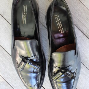 May include: A pair of black leather tassel loafers with the brand name "Johnston & Murphy" embossed on the inside of each shoe.