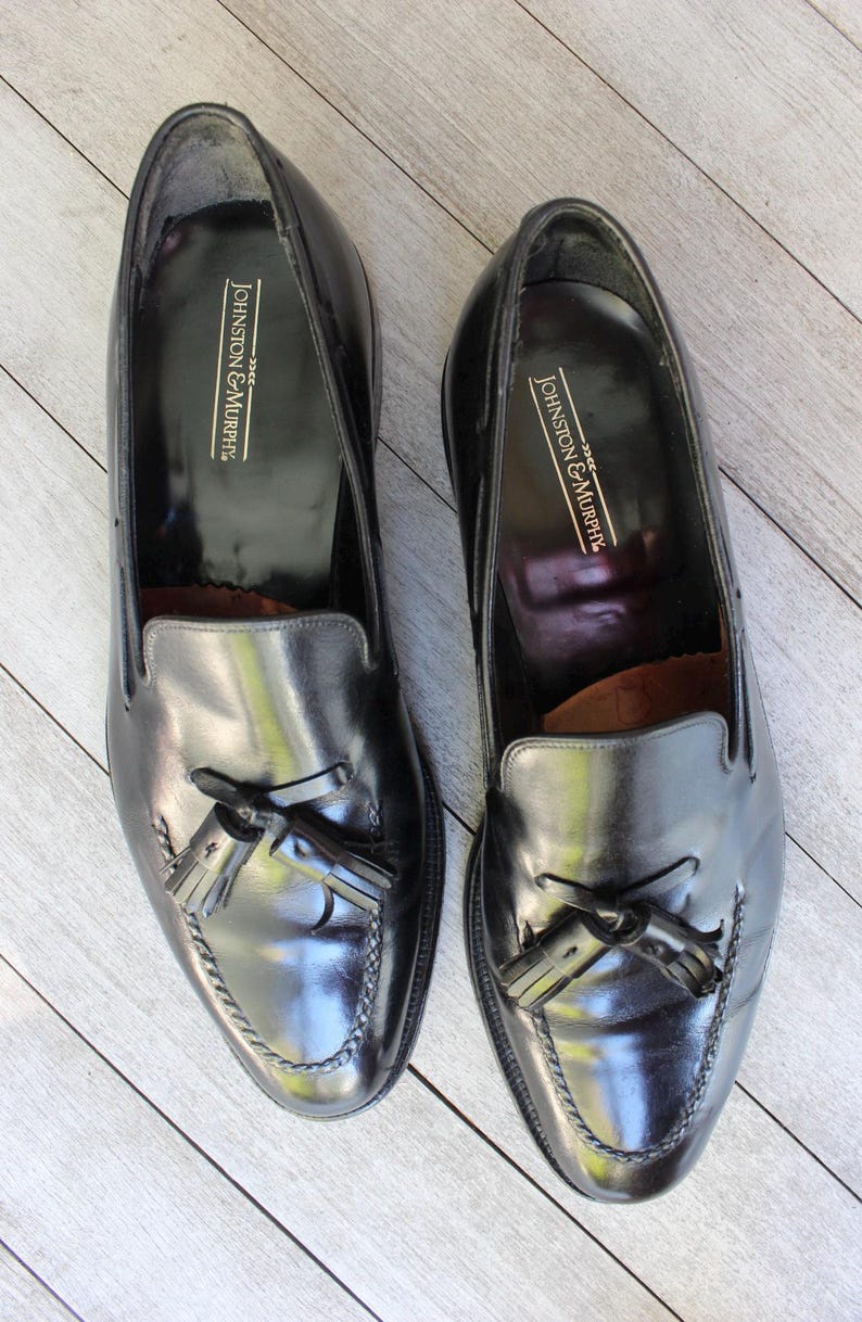 May include: A pair of black leather tassel loafers with the brand name "Johnston & Murphy" embossed on the inside of each shoe.
