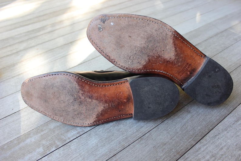 May include: Close-up of the soles of a pair of black leather shoes. The shoes have a brown leather sole and a black rubber heel.
