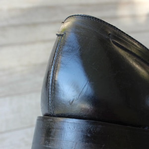 May include: Close-up of the back of a black leather dress shoe with a stitched seam. The shoe has a thick sole and a rounded toe.