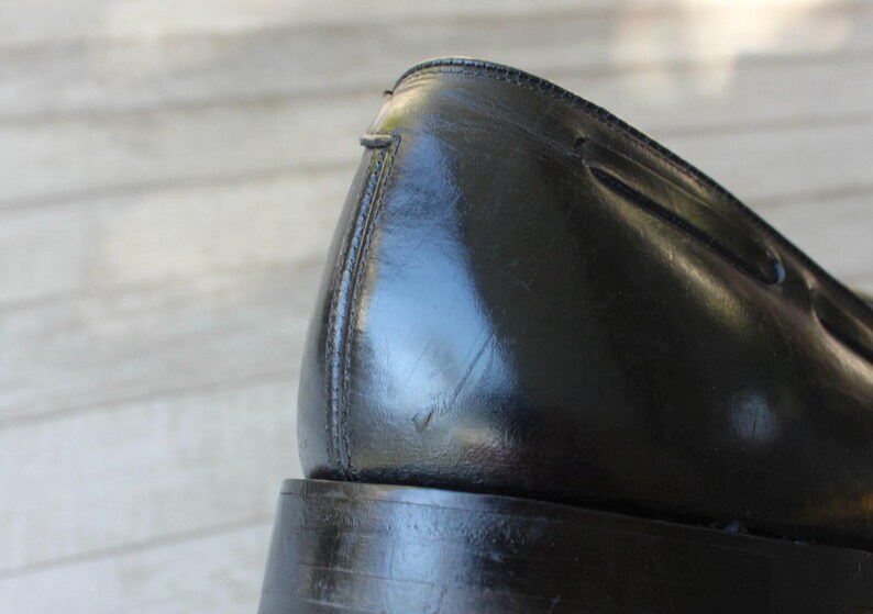 May include: Close-up of the back of a black leather dress shoe with a stitched seam. The shoe has a thick sole and a rounded toe.