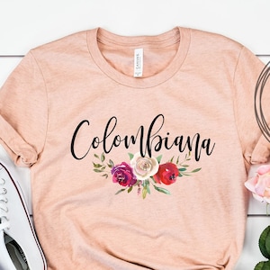 May include: A peach-colored t-shirt with the text "Colombiana" in black script with a floral design underneath. The shirt is laid out on a white wooden surface with a pair of white sneakers and a bouquet of pink roses.