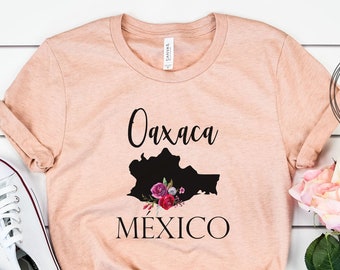 May include: A peach-coloured t-shirt with a black silhouette of the state of Oaxaca, Mexico, with pink and red floral accents. The text "Oaxaca" and "Mexico" are printed in black.