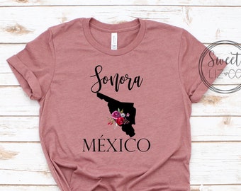May include: A mauve t-shirt featuring a black outline of the state of Sonora, Mexico, with the text "Sonora" and "MÉXICO" printed on the shirt. The outline of the state is decorated with pink and purple flowers.
