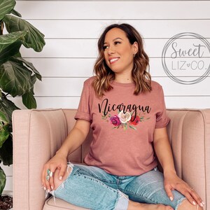 May include: A mauve t-shirt with a floral design and the word "Nicaragua" printed in black. The shirt is being worn by a person sitting on a pink couch.