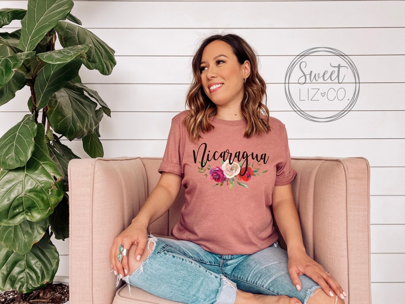 May include: A mauve t-shirt with a floral design and the word "Nicaragua" printed in black. The shirt is being worn by a person sitting on a pink couch.