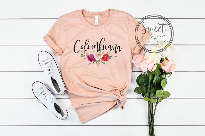 May include: A peach-colored t-shirt with the text "Colombiana" in black script with a floral design underneath. The shirt is laid out on a white wooden surface with a pair of white sneakers and a bouquet of pink roses.