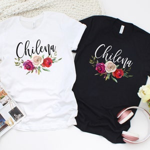 May include: Two t-shirts with the word "Chilena" in a cursive font and a floral design with red, pink, and white flowers. The t-shirts are one white and one black.
