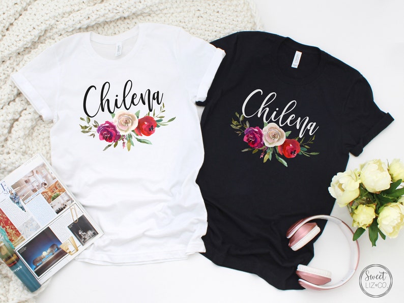 May include: Two t-shirts with the word "Chilena" in a cursive font and a floral design with red, pink, and white flowers. The t-shirts are one white and one black.