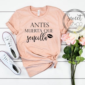May include: A peach-colored t-shirt with the text "Antes Muerta Que Sencilla" and a black lip print. The shirt is laid flat on a white wooden surface with a bouquet of pink roses and white sneakers.