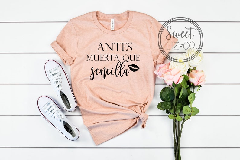 May include: A peach-colored t-shirt with the text "Antes Muerta Que Sencilla" and a black lip print. The shirt is laid flat on a white wooden surface with a bouquet of pink roses and white sneakers.