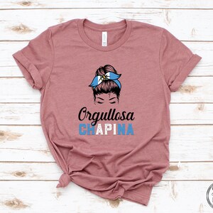 Puede incluir: Una camiseta color malva con un gr&aacute;fico de una mujer con un mo&ntilde;o en el pelo que lleva una diadema azul y blanca con el texto "Orgullosa CHAPINA" debajo.