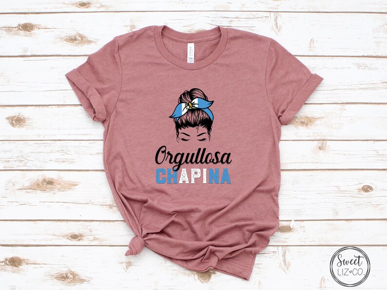 Puede incluir: Una camiseta color malva con un gr&aacute;fico de una mujer con un mo&ntilde;o en el pelo que lleva una diadema azul y blanca con el texto "Orgullosa CHAPINA" debajo.