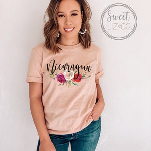 May include: A light pink t-shirt with the word "Nicaragua" printed in black script. The shirt features a floral design with pink, red, and white flowers.