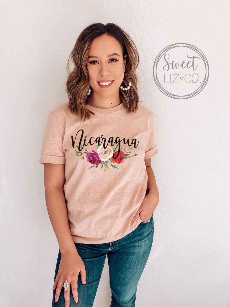 May include: A light pink t-shirt with the word "Nicaragua" printed in black script. The shirt features a floral design with pink, red, and white flowers.