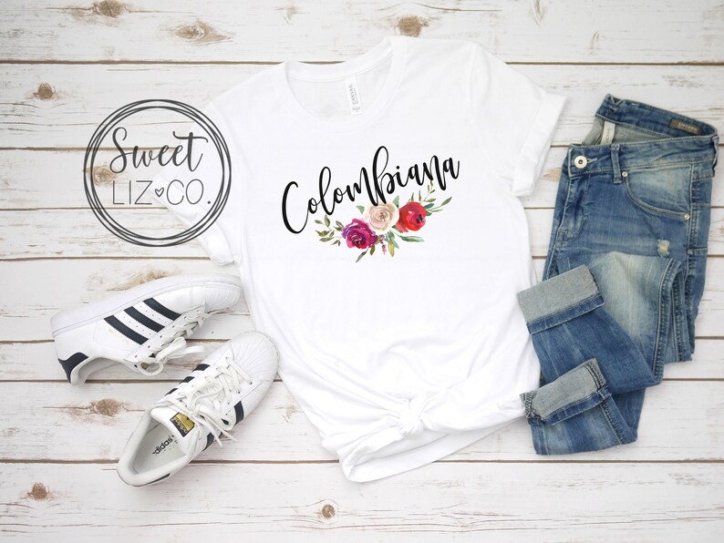 May include: White t-shirt with the text "Colombiana" in black script with a floral design in pink, red, and white.