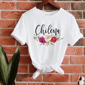 May include: White t-shirt with the word "Chilena" written in black cursive script and a floral design with red, pink, and white flowers.