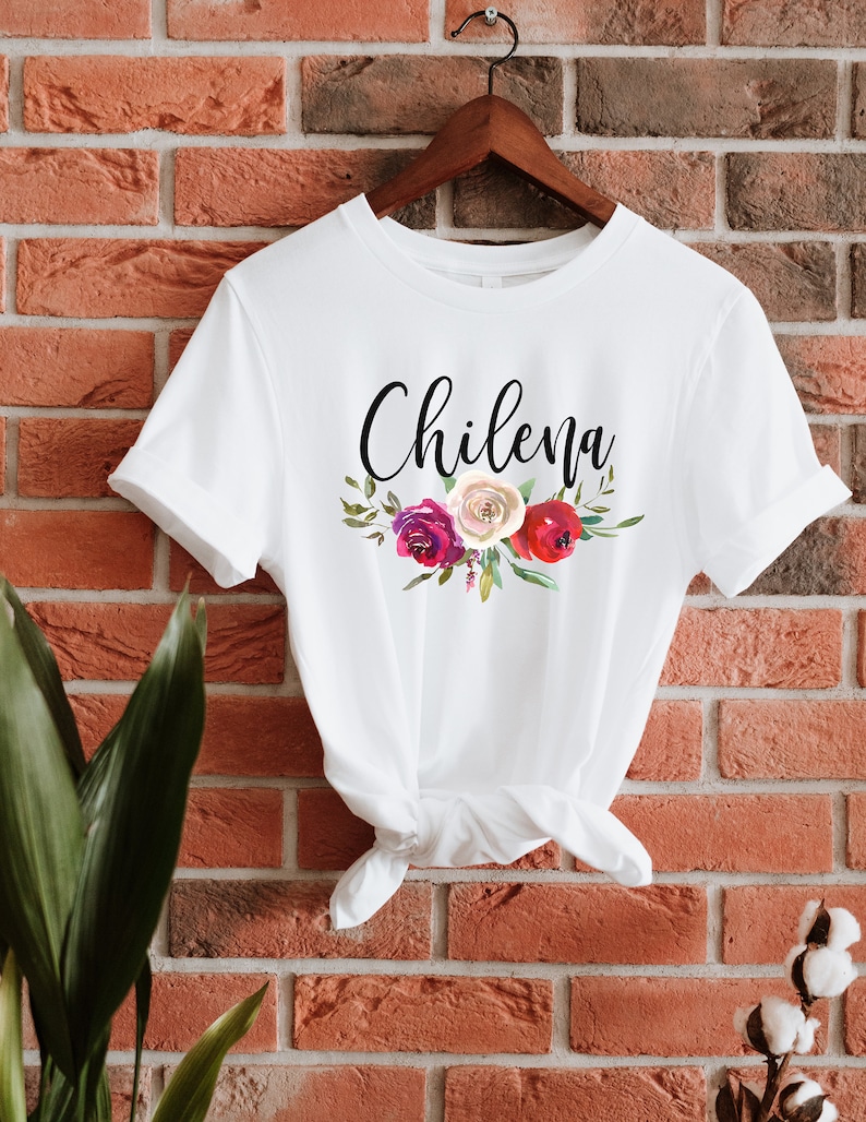 May include: White t-shirt with the word "Chilena" written in black cursive script and a floral design with red, pink, and white flowers.