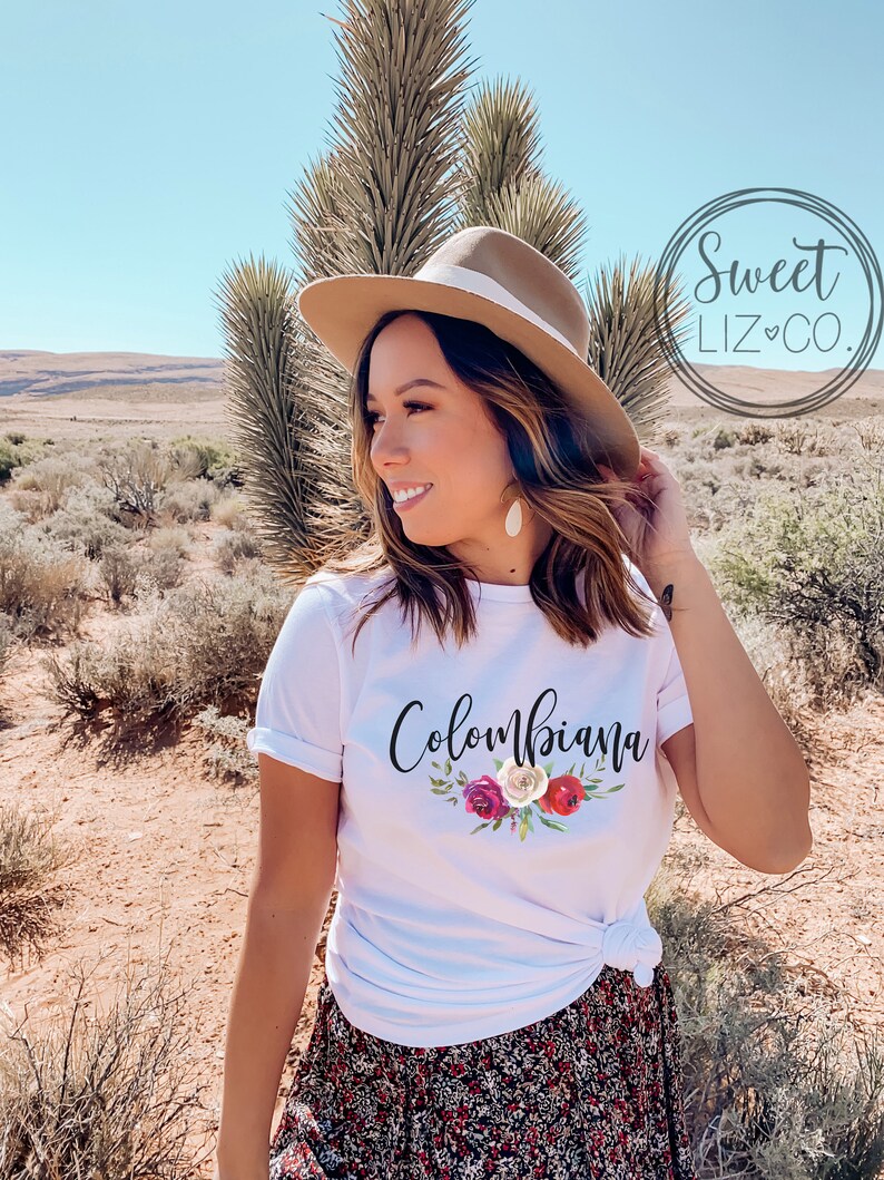 May include: White t-shirt with the text "Colombiana" in black script with a floral design in red, white, and pink.